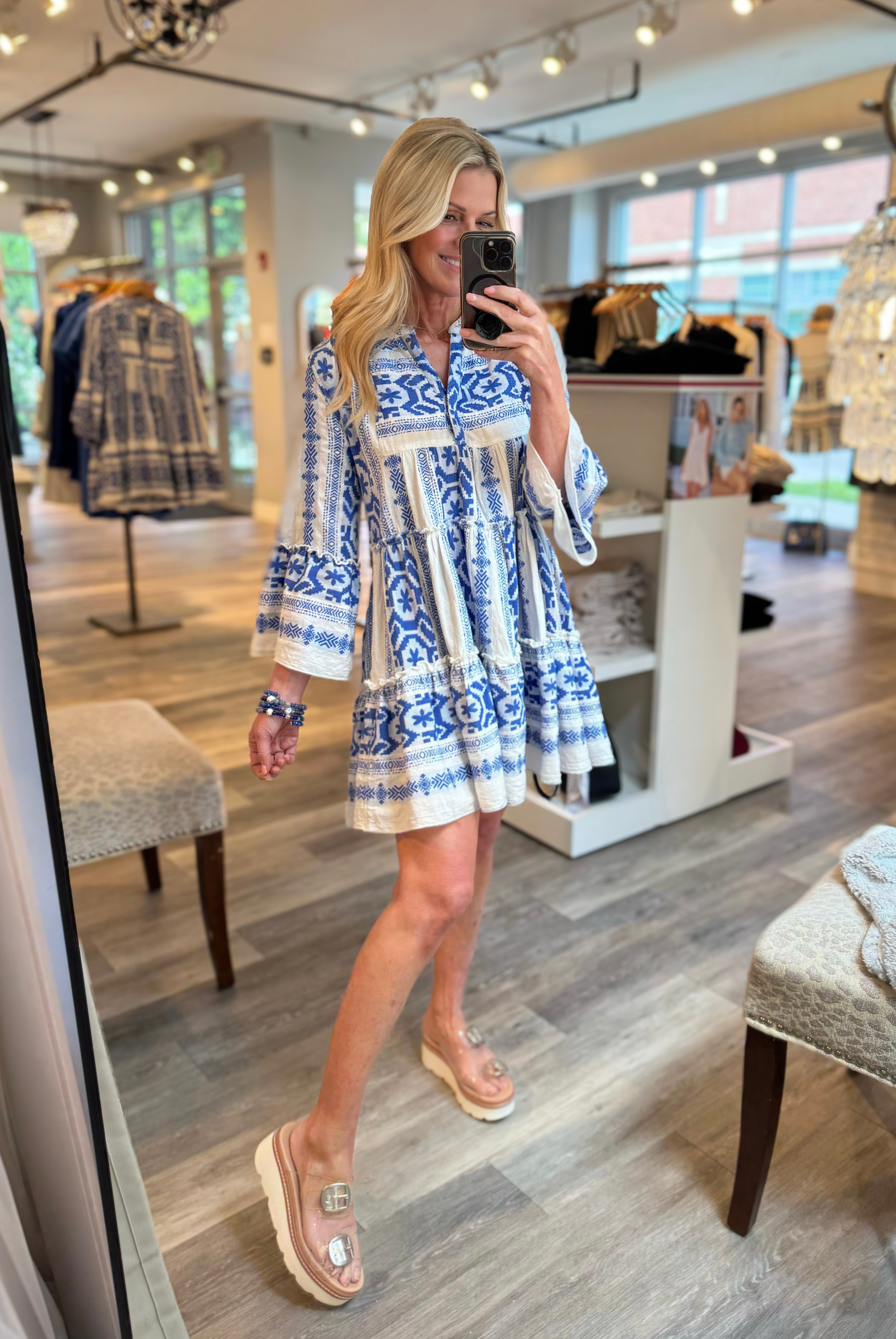 A woman takes a mirror selfie in a boutique while wearing a blue and white patterned mini dress with bell sleeves and clear sandals.