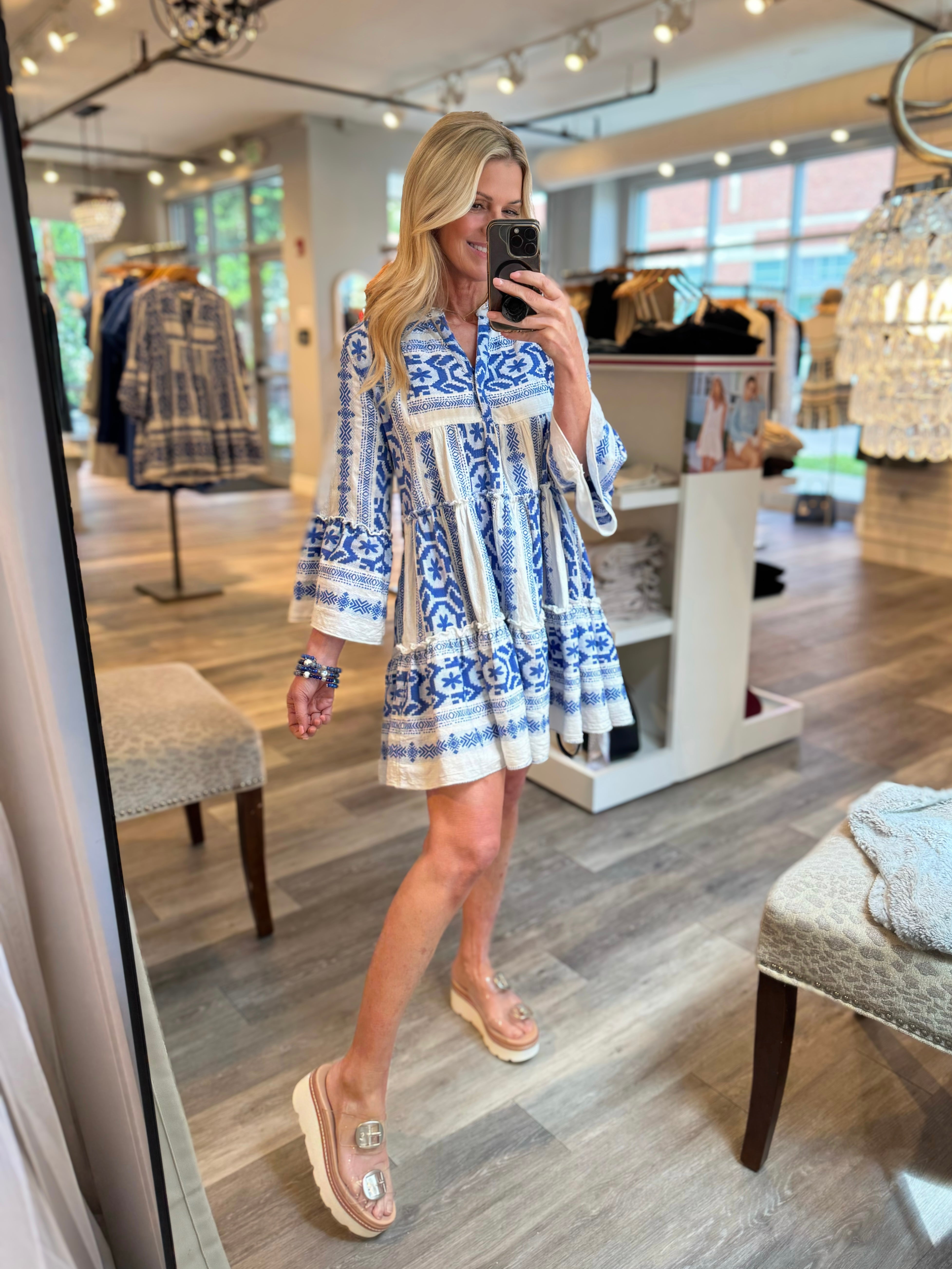 A woman takes a mirror selfie in a boutique while wearing a blue and white patterned mini dress with bell sleeves and clear sandals.
