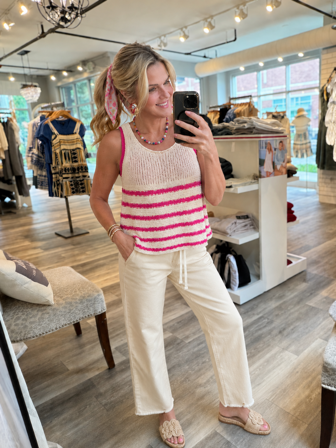 A woman takes a mirror selfie in a clothing boutique, wearing a cream knit tank top with bright pink horizontal stripes and cream wide-leg drawstring pants.
