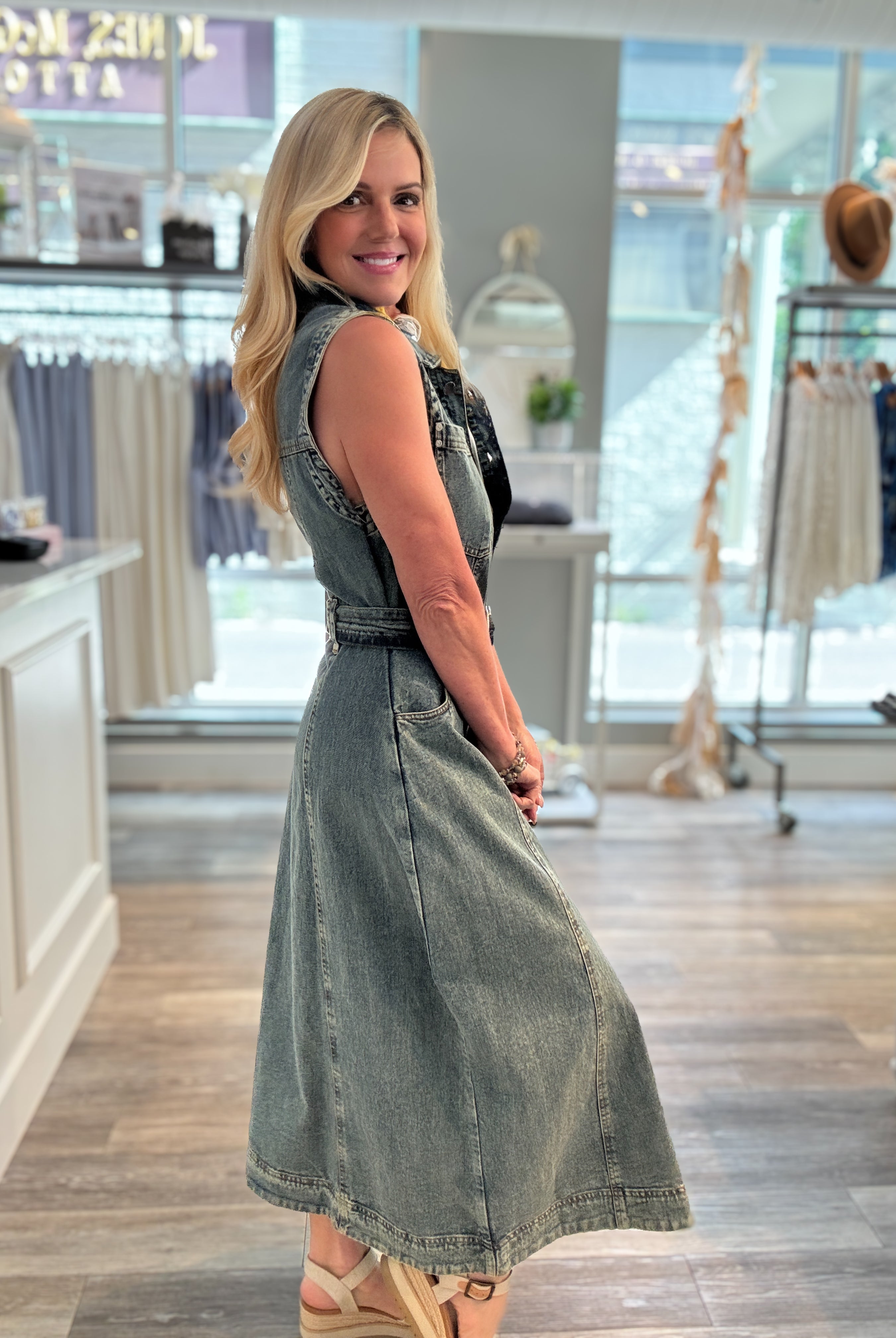 A woman modeling side of a sleeveless denim midi dress with a belted waist inside a boutique clothing store.