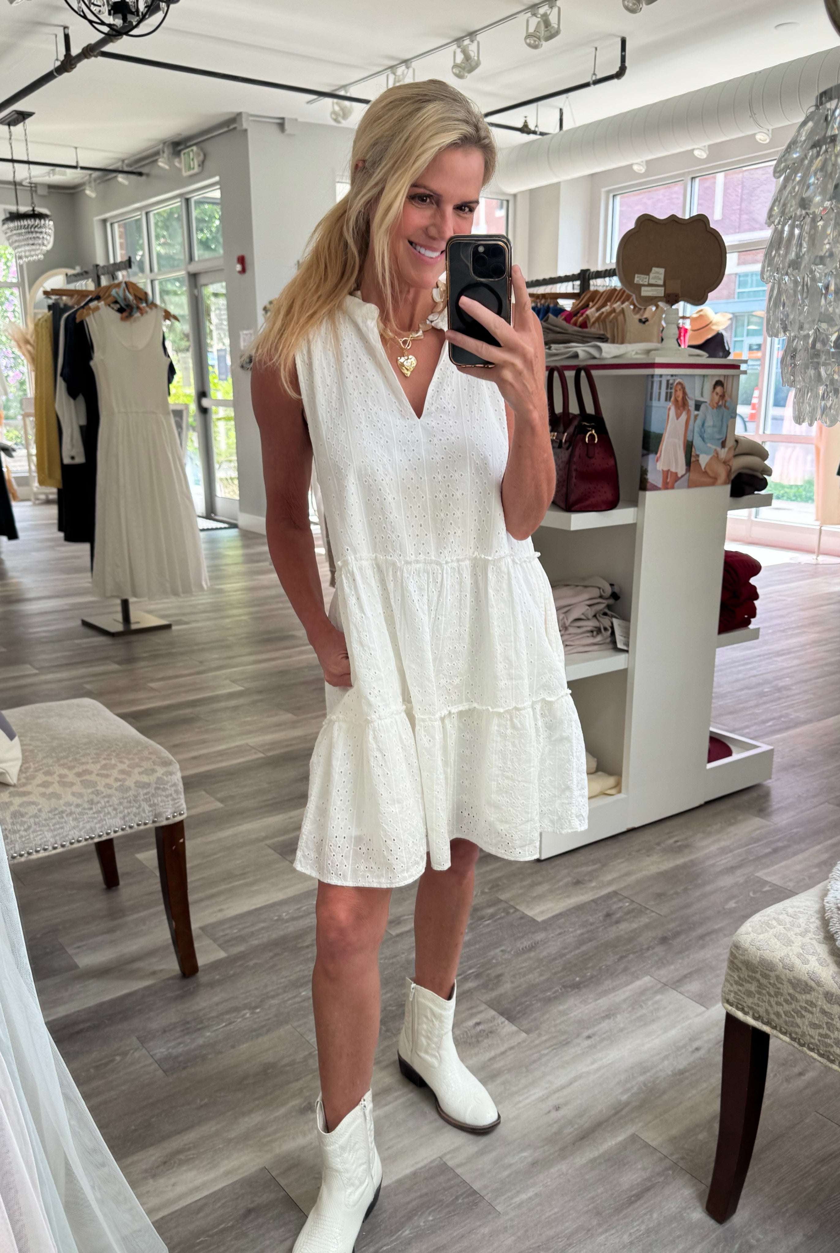 A woman takes a mirror selfie in a boutique wearing a sleeveless white tiered mini dress and white cowboy boots.