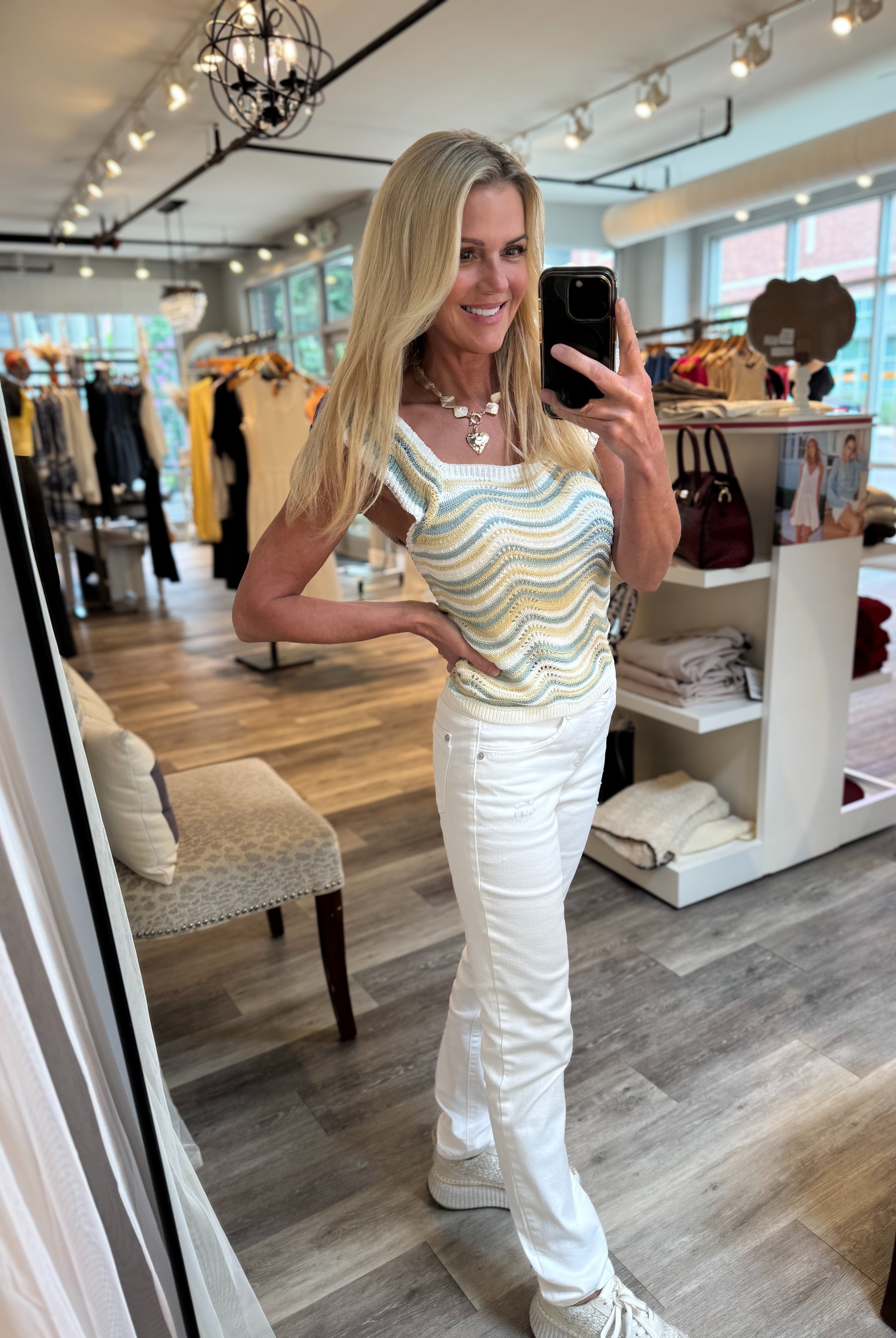 A woman takes a mirror selfie in a clothing boutique, wearing a sleeveless wavy striped tank top in pastel yellow, green, and blue, paired with white pants and white sneakers.