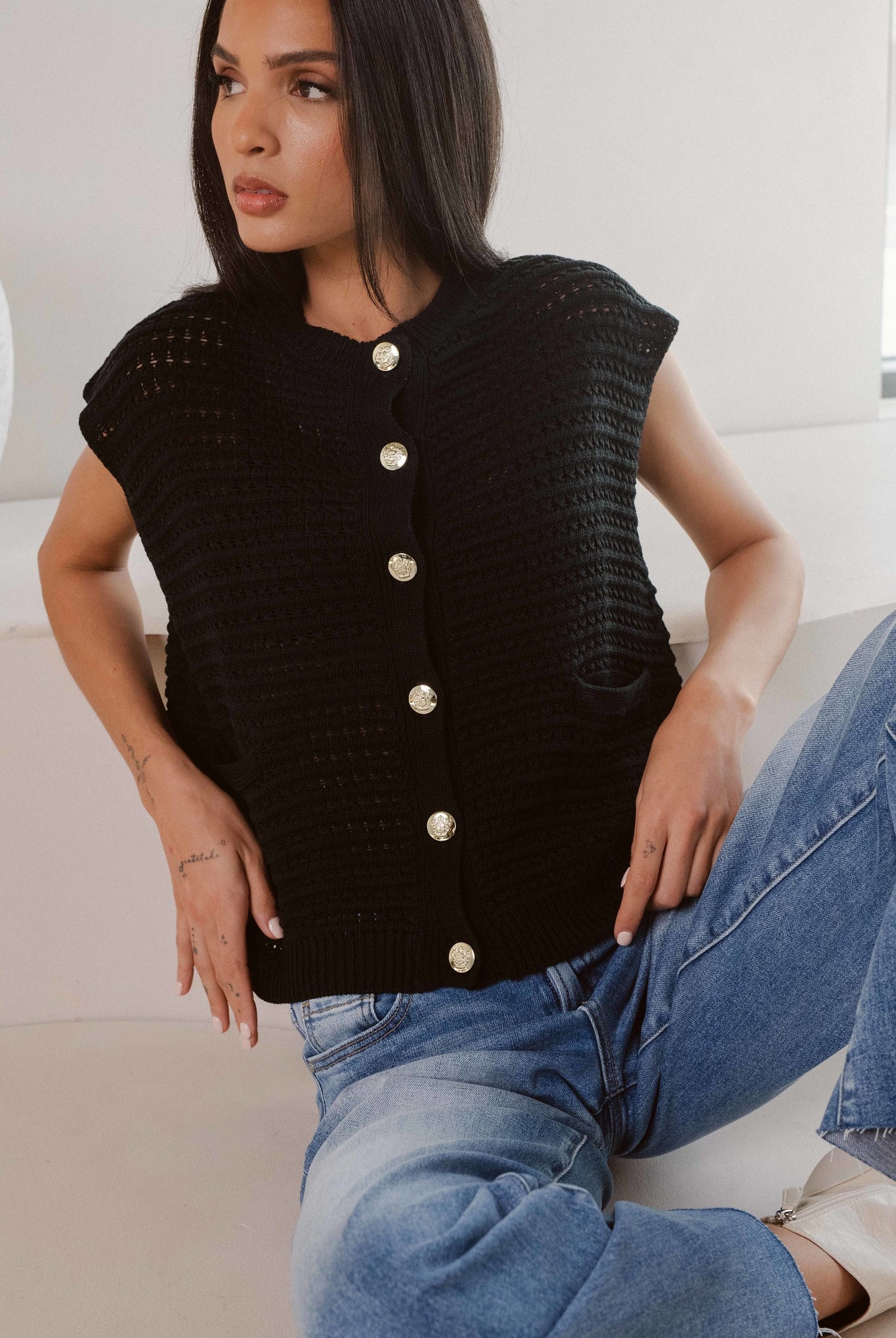 Model wearing black knitted cap sleeve vest with silver buttons, styled with denim jeans