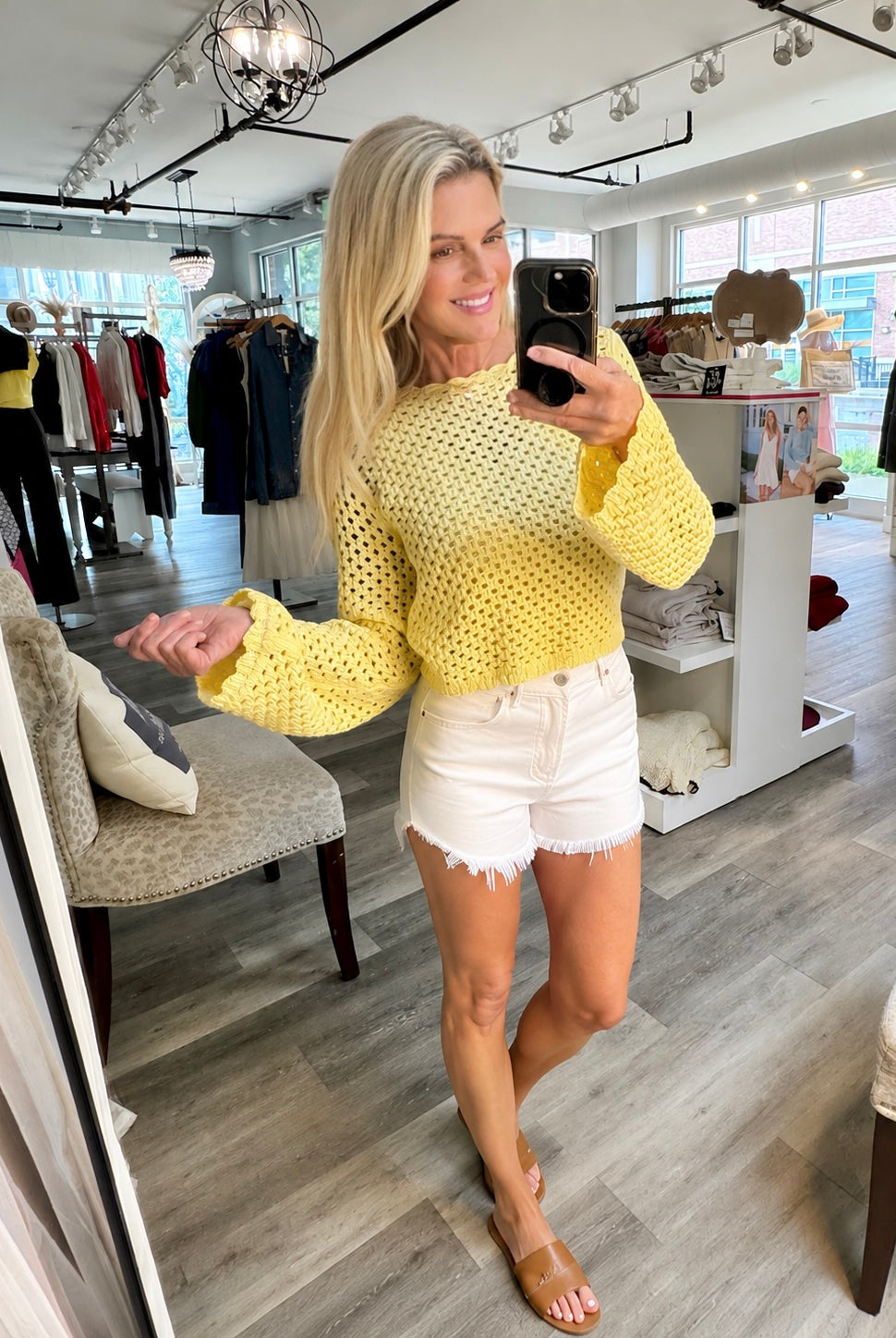 A woman takes a mirror selfie in a clothing boutique wearing a butter yellow open-knit long-sleeve sweater white denim shorts, and tan flat sandals.
