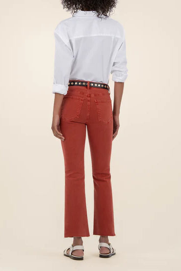 Woman showing the back of the high rise ankle flare jeans in the color paprika along with a white button down shirt and a black belt.