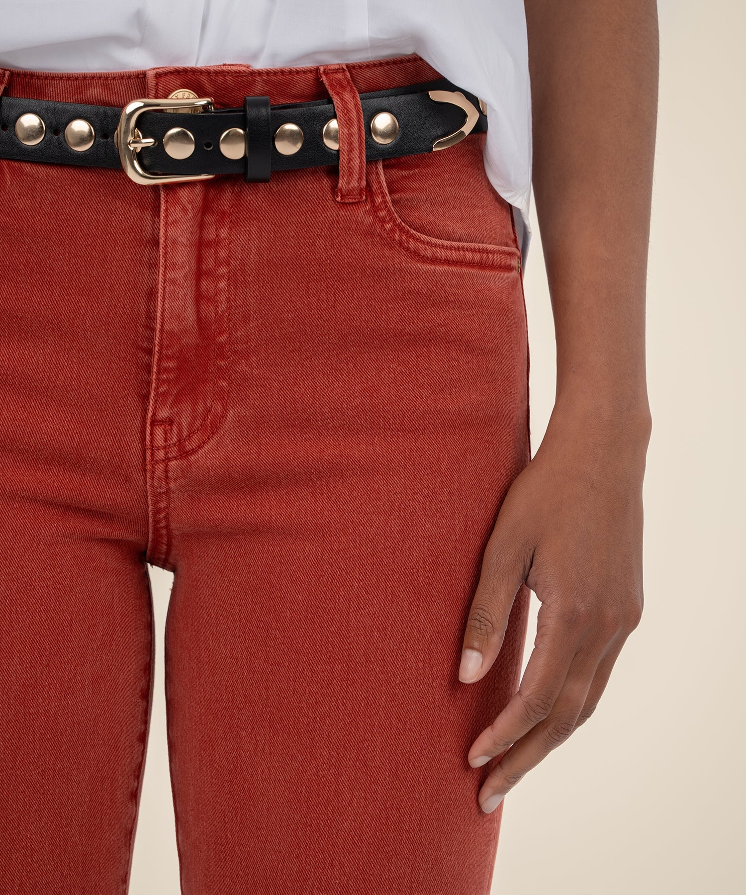 Woman showing the details of the high rise ankle flare jeans in the color paprika along with a white button down shirt and a black belt.
