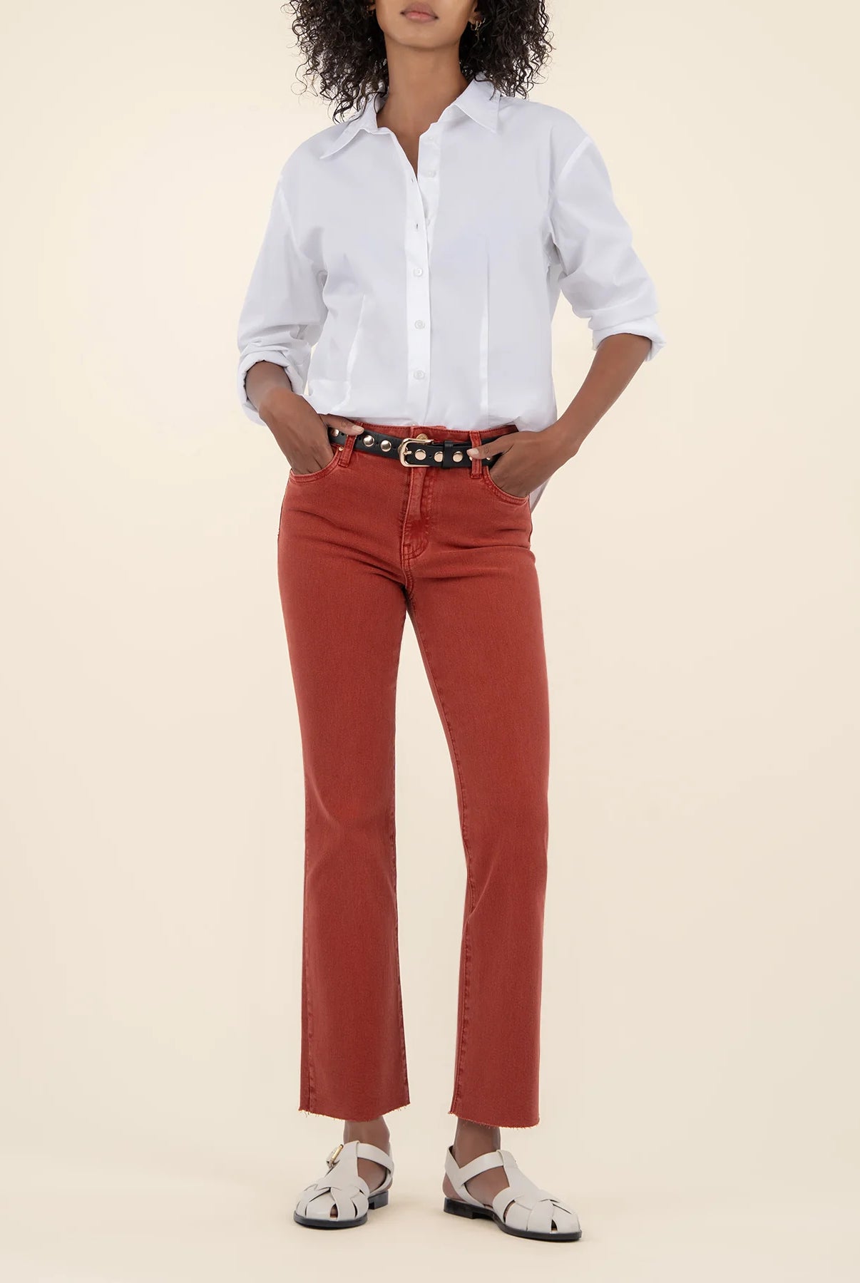 Woman wearing the high rise ankle flare jeans in the color paprika along with a white button down shirt and a black belt.