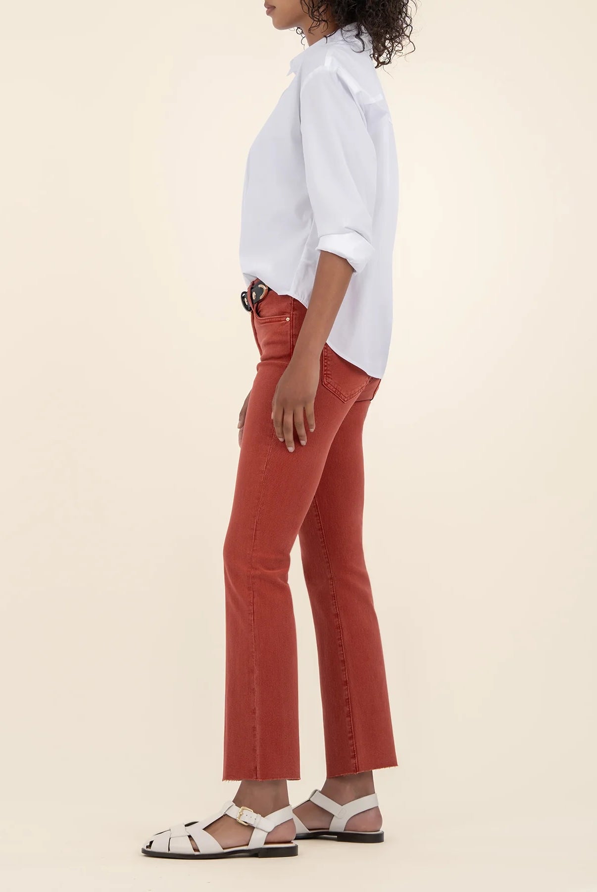 Woman showing the side of the high rise ankle flare jeans in the color paprika along with a white button down shirt and a black belt.