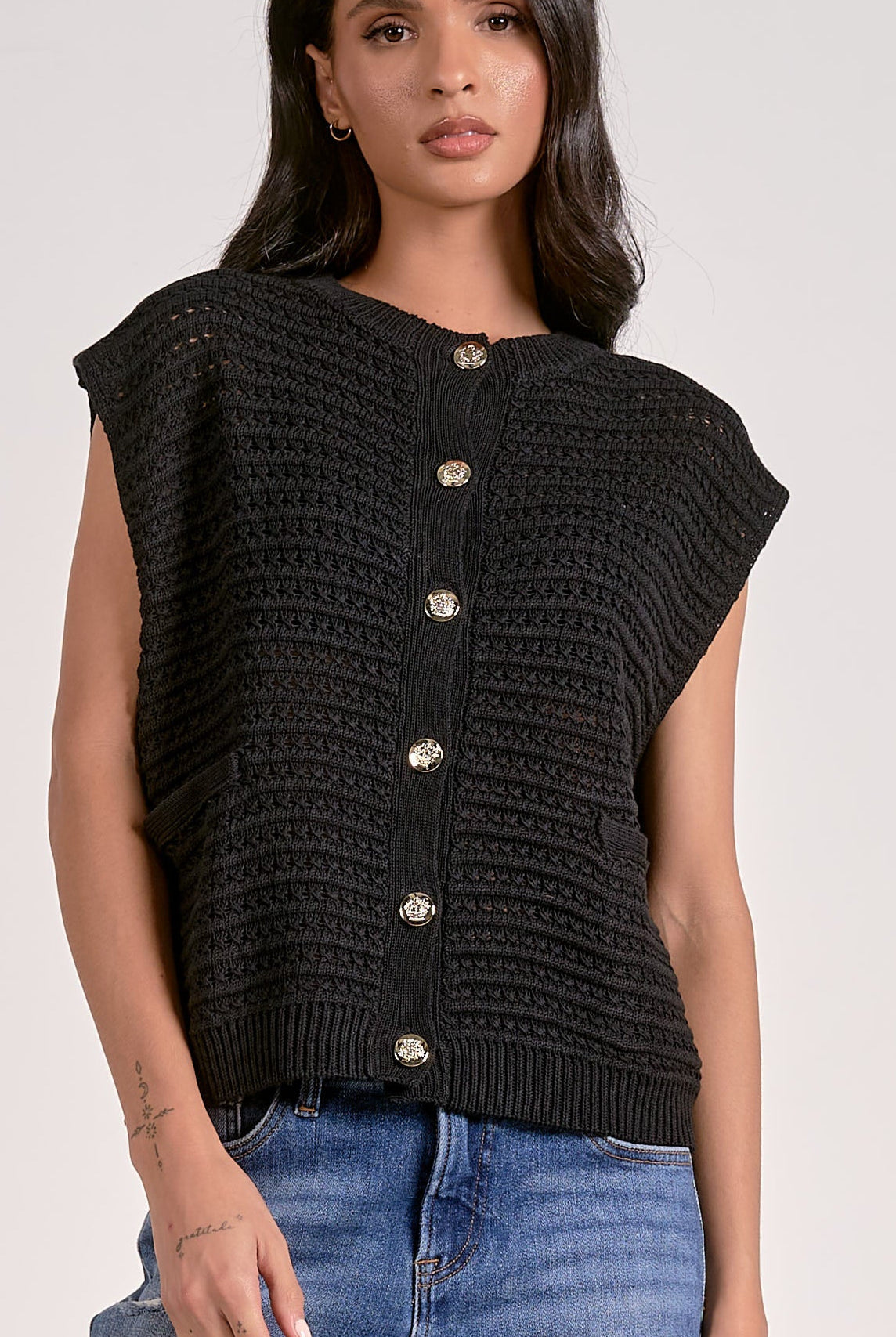 Model wearing black knitted cap sleeve vest with silver buttons, styled with denim jeans