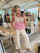 A woman takes a mirror selfie in a clothing boutique, wearing a cream knit tank top with bright pink horizontal stripes and cream wide-leg drawstring pants.
