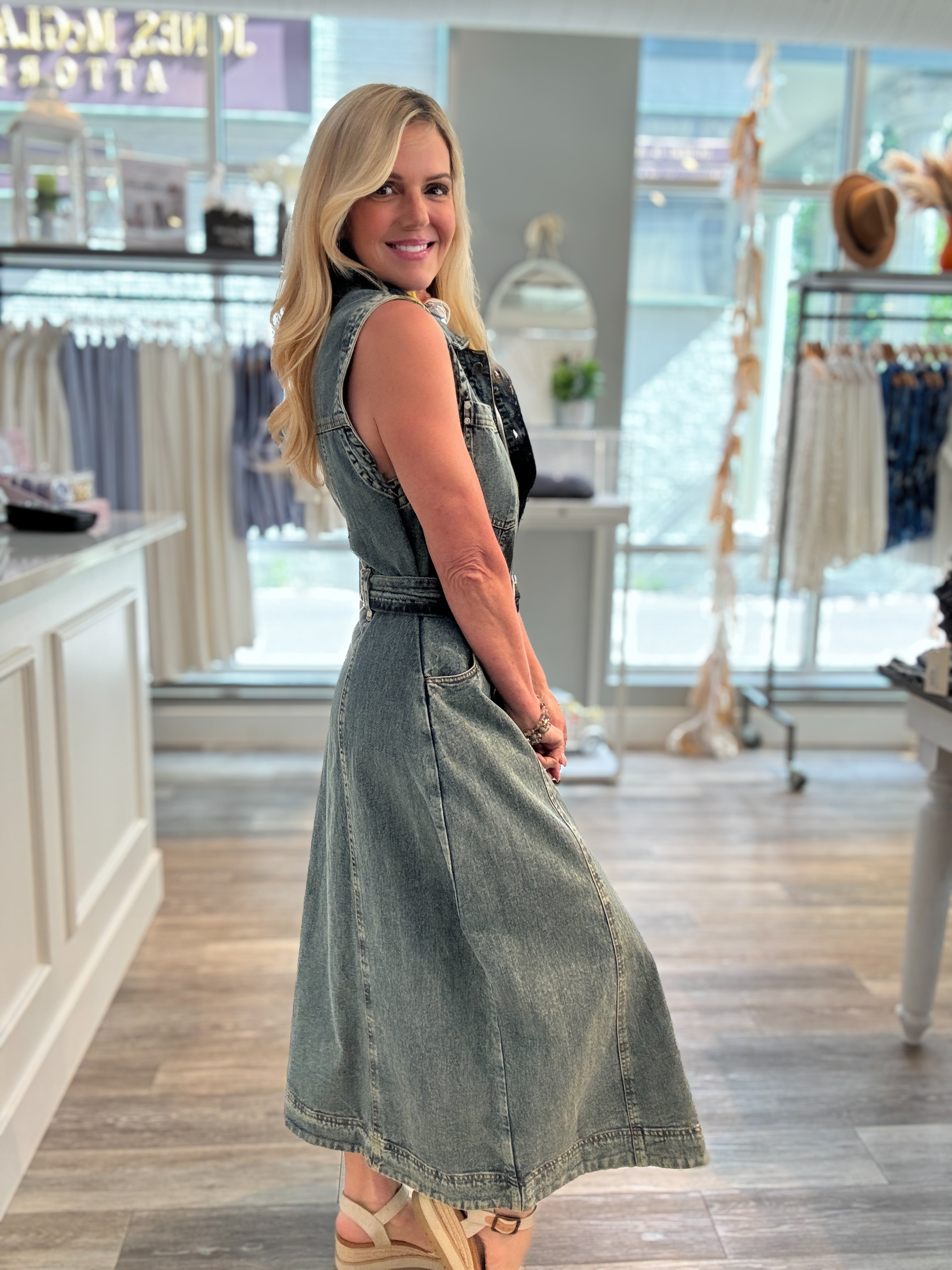 A woman modeling side of a sleeveless denim midi dress with a belted waist inside a boutique clothing store.