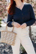 Person wearing a Pointelle Cardigan with Rose Button and white pants, holding a woven bag with blurred background
