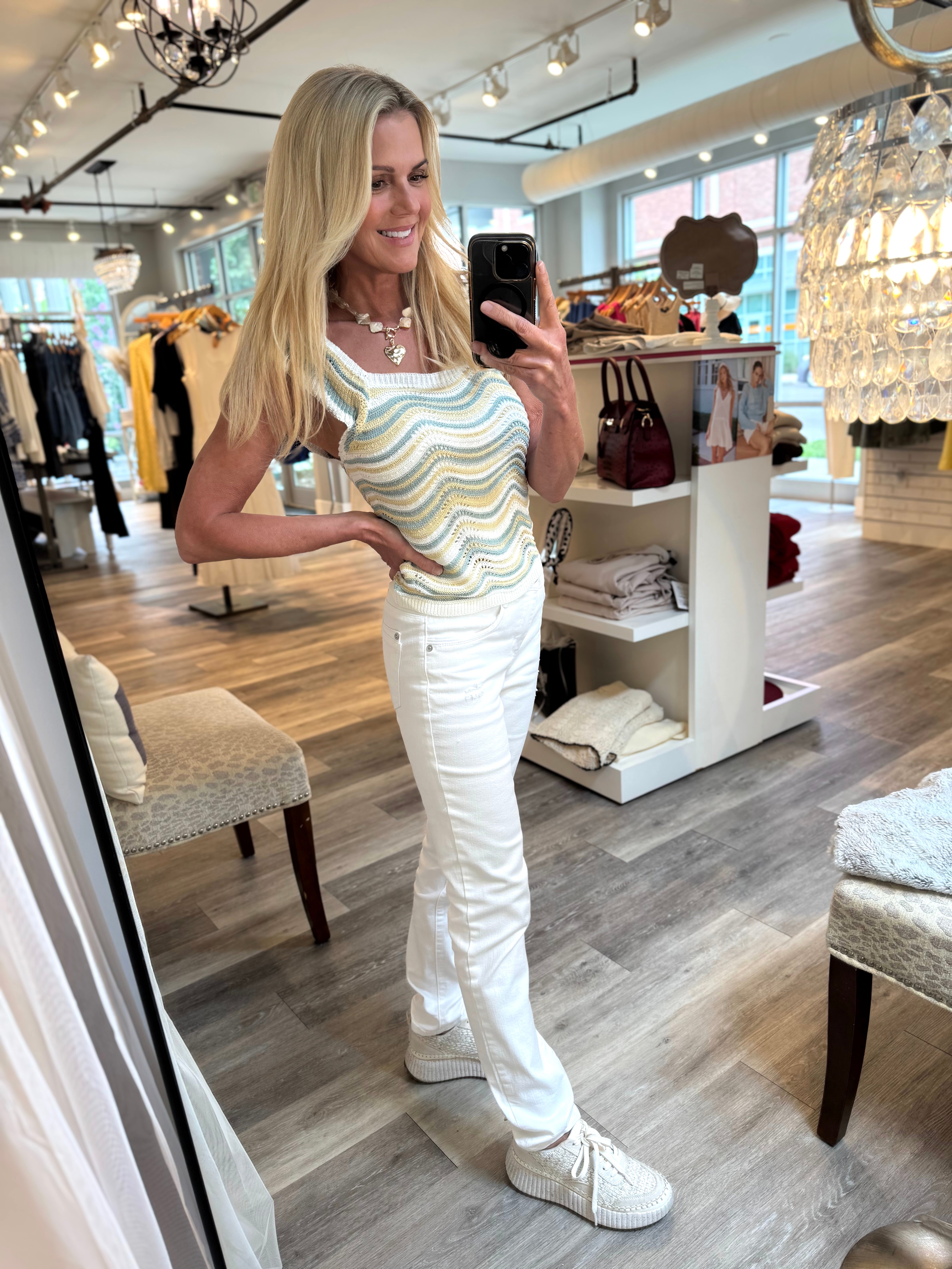 A woman takes a mirror selfie in a boutique, wearing a sleeveless top with a wavy multicolor stripe pattern in green, yellow, and white, paired with white pants and white sneakers.