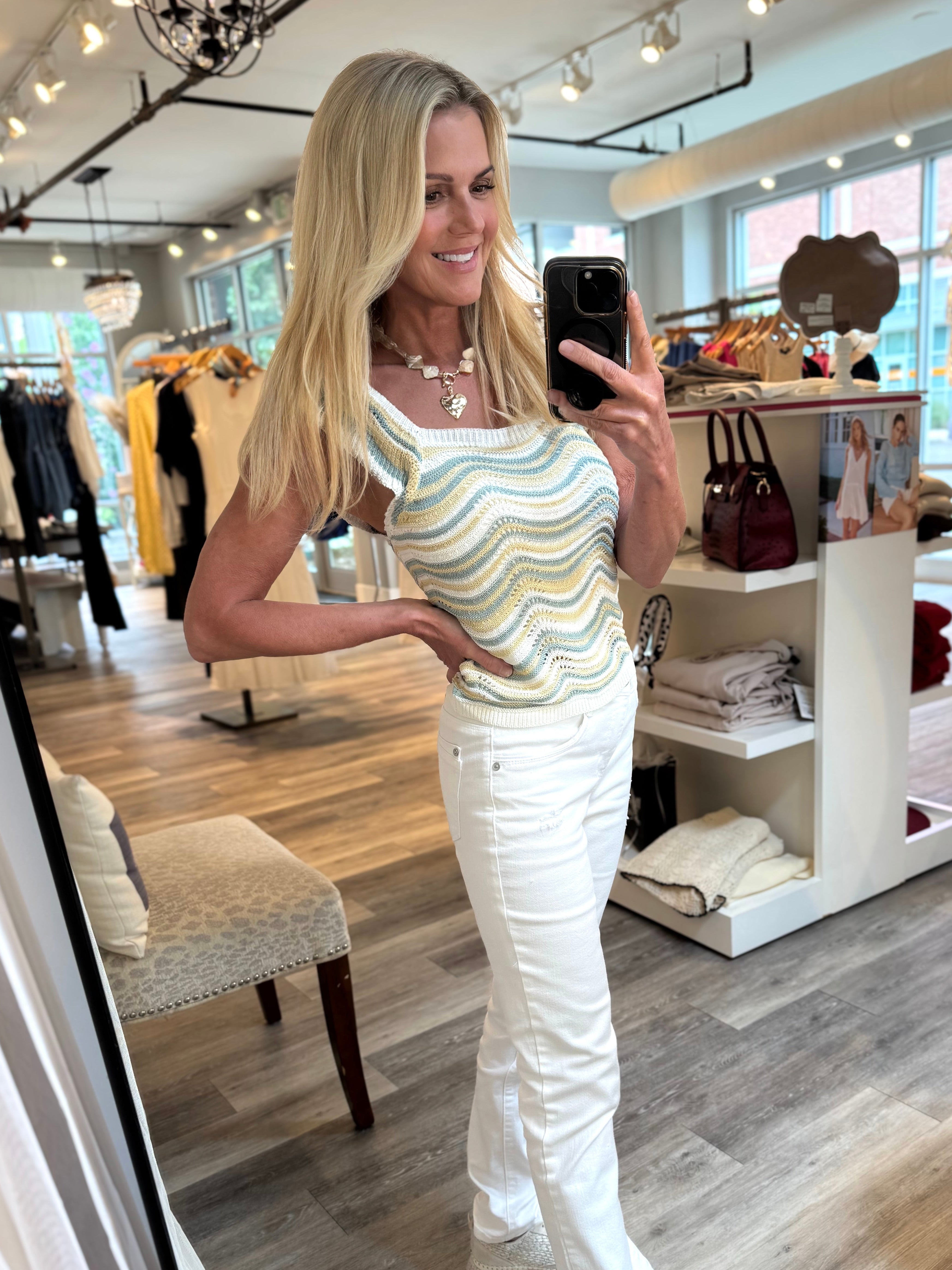 A woman takes a mirror selfie in a boutique, wearing a sleeveless top with a wavy multicolor stripe pattern in green, yellow, and white, paired with white pants and white sneakers.