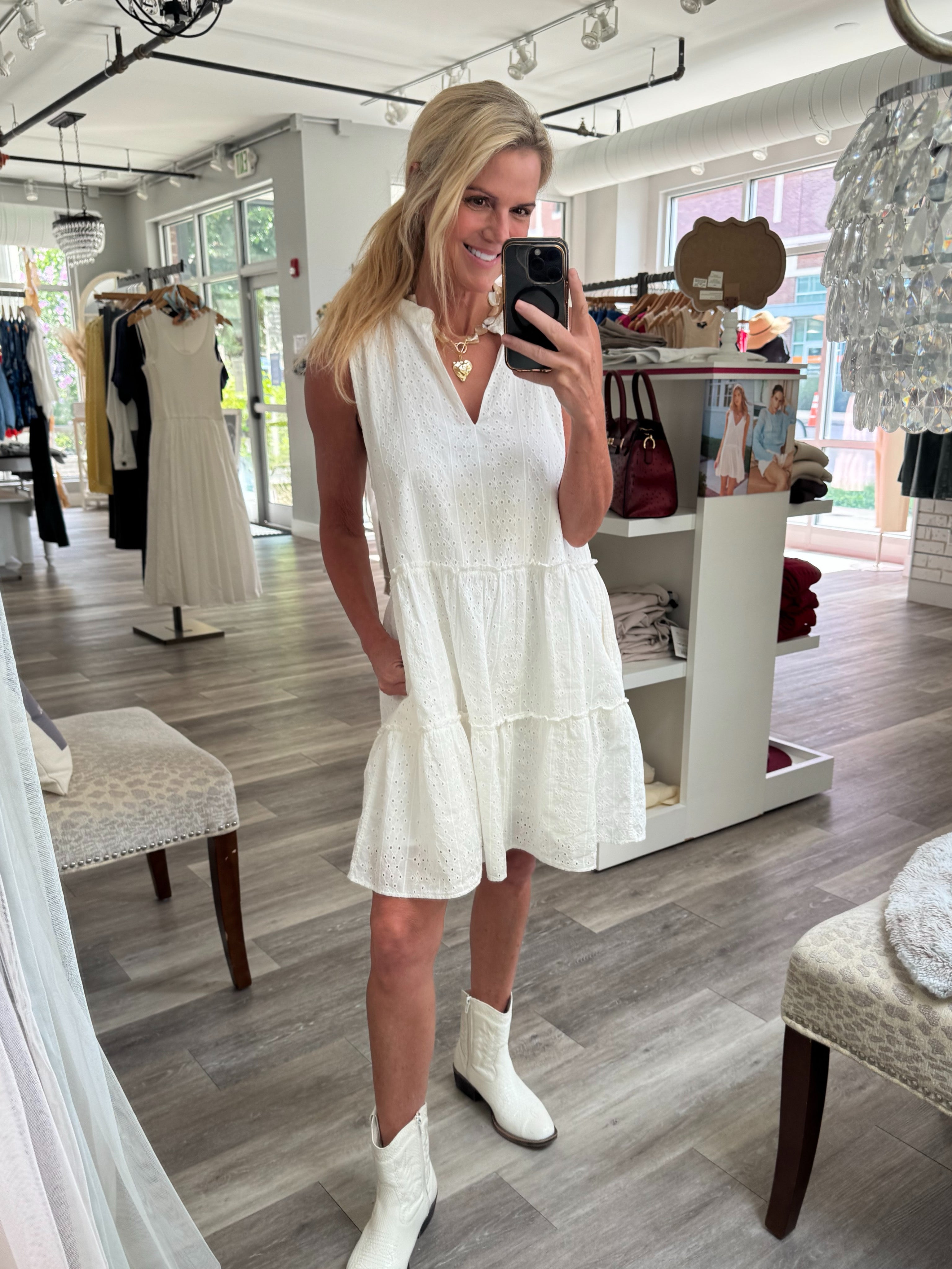 A woman takes a mirror selfie in a boutique wearing a sleeveless white tiered mini dress and white cowboy boots.