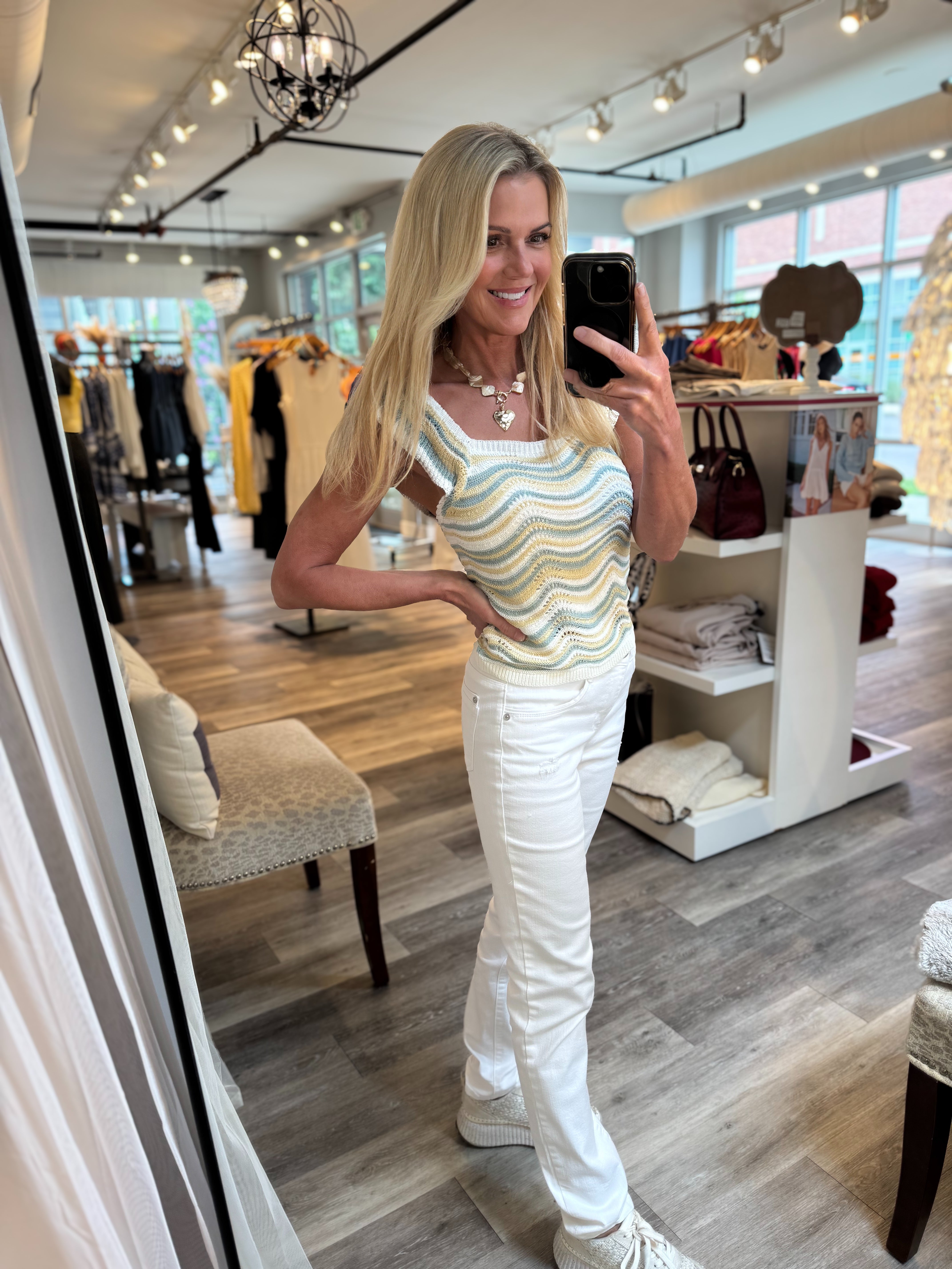 A woman takes a mirror selfie in a clothing boutique, wearing a sleeveless wavy striped tank top in pastel yellow, green, and blue, paired with white pants and white sneakers.