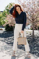Woman in Pointelle Cardigan with Rose Button and white pants holding a woven bag outdoors with cherry blossoms in the background.
