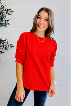 Woman wearing a red top and blue jeans standing against a white wall with a plant to the left.