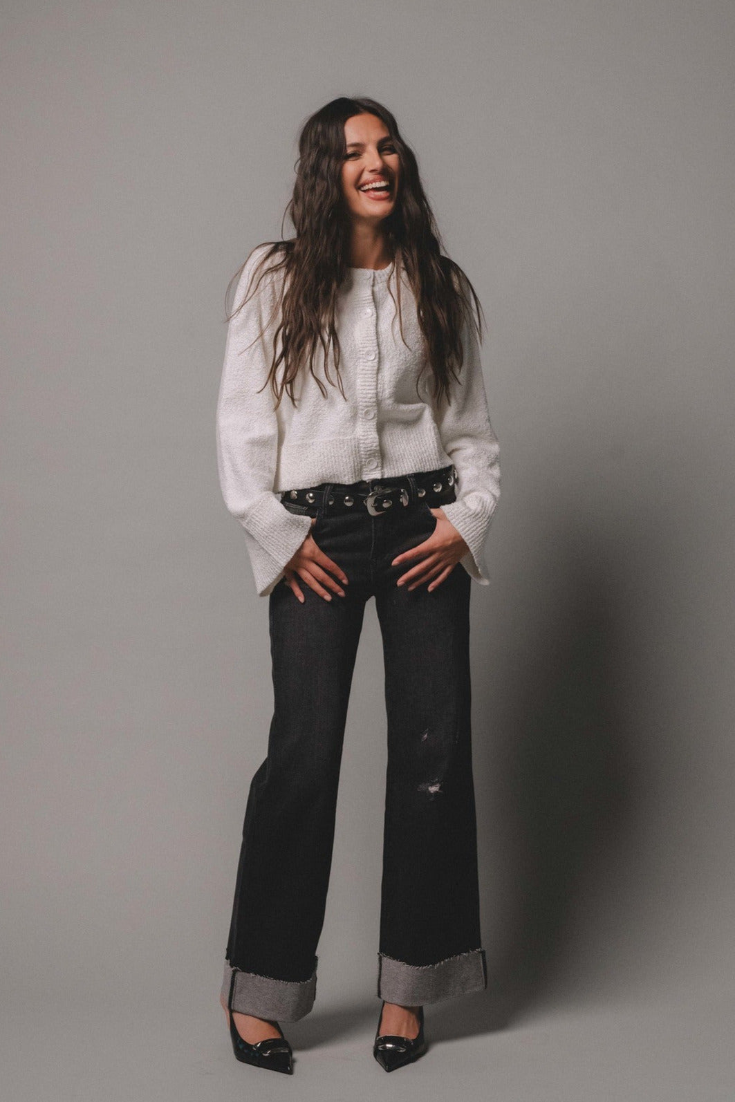 Woman completing the look of the ivory button down cardigan with a belt and cuff bottom jeans.