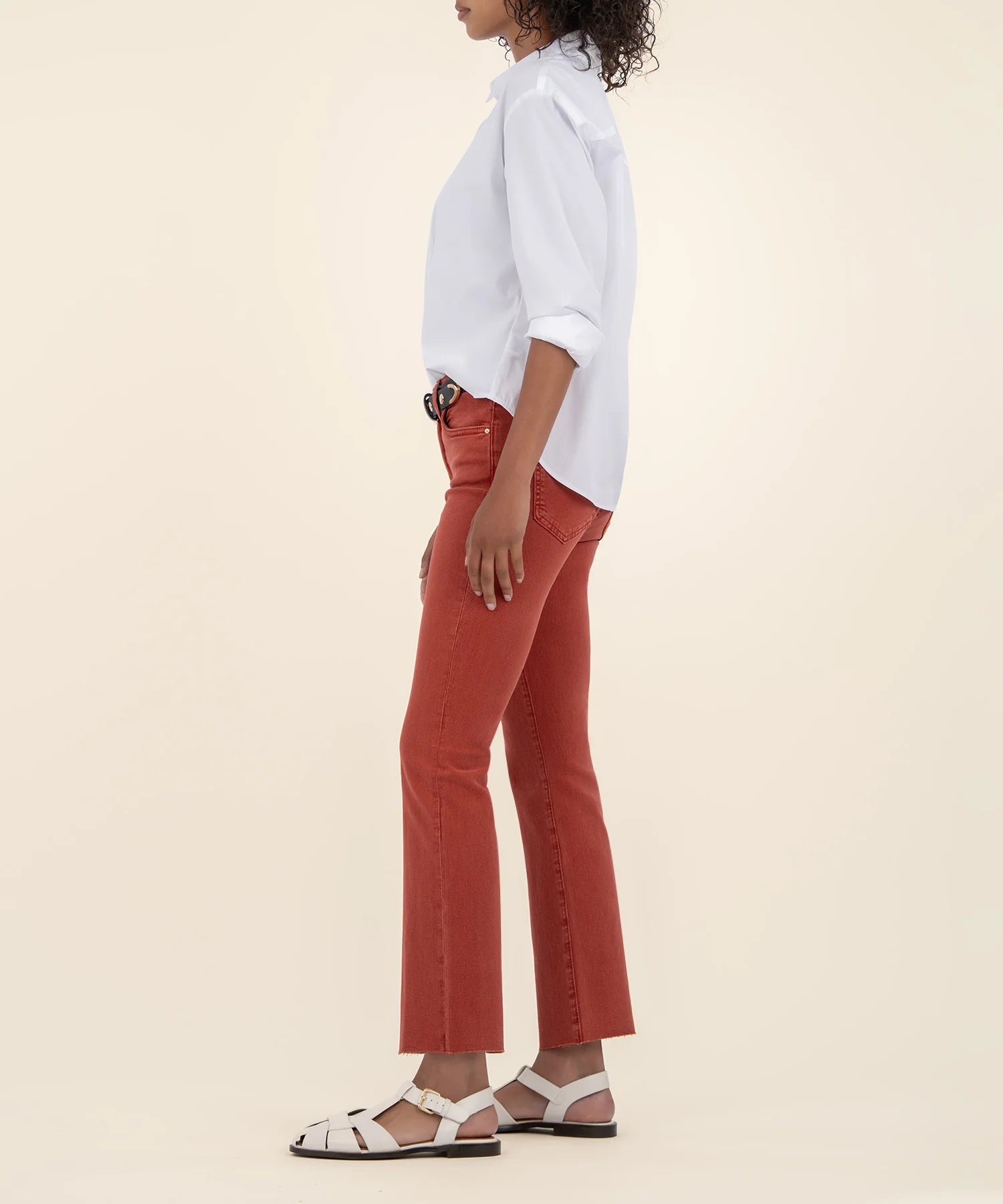 Woman showing the side of the high rise ankle flare jeans in the color paprika along with a white button down shirt and a black belt.